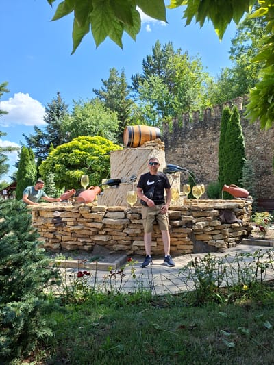 In Milestii Mici, Moldova, a man stands posing in front of a decorative fountain that mimics a wine barrel, glasses, and amphorae. The fountain is constructed from stacked rough-hewn stones and is situated amidst lush greenery, including pine trees and flowering bushes. A stone wall forms the backdrop. Another man is visible in the background, leaning over the fountain. The scene is bathed in sunlight under a clear blue sky.