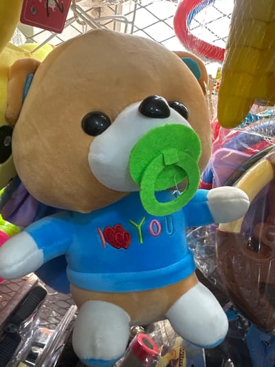 A plush teddy bear toy, approximately 2-3 feet tall, is the central subject of the image. The bear has light brown fur, black button eyes, and a white muzzle with a green pacifier. It is wearing a blue long-sleeved shirt with the embroidered text "I LOVE YOU" in red and pink. The bear is positioned in a store, likely a toy shop, with shelves filled with other toys and merchandise visible in the background. The lighting appears to be artificial, casting soft shadows. The overall impression is of a retail environment displaying children's toys. The image is taken from a medium-low angle, focusing on the bear. The environment suggests a consumer setting where such toys are sold.
