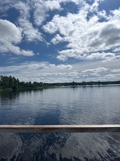 A wide, calm lake with ripples is in the foreground, reflecting the sky. The far bank is lined with a dense forest of green trees. Above the trees, a bright blue sky is filled with fluffy white clouds, some of which are large and voluminous, while others are wispy and scattered. A rusty metal railing runs horizontally across the bottom of the image, obscuring the lower portion of the lake and its reflections. The overall scene depicts a serene natural landscape on a clear, sunny day. The image was captured in Vazhiny, Russia.