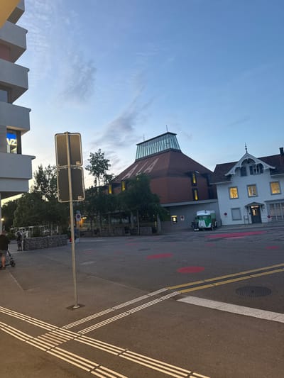 This media file captures an evening street scene in Liestal, Switzerland, rotated 90 degrees clockwise from its natural orientation. The sky, seen on the left (top when correctly oriented), is a clear, light blue with faint wispy clouds, indicating either dusk or early evening, a time reinforced by the illuminated windows in several buildings.
The setting is an urban street flanked by diverse architecture. On the far right (top when correctly oriented), stands a charming, traditional white building with a patterned dark roof and decorative gables, its windows glowing with warm light. Adjacent to it, closer to the center, is a larger building featuring a reddish-brown tiled roof and a distinctive modern glass and metal section, likely a skylight or roof extension. This building also has illuminated windows. Further to the left (bottom when correctly oriented), modern architectural elements with sharp, angular designs are visible. One such structure displays glowing blue letters, possibly "VZ" or "VZA," indicating text on a sign or display.
The paved asphalt road occupies a significant portion of the foreground and center. It features clear road markings, including double solid yellow lines and distinct triple white lines, possibly indicating a pedestrian crossing or a specific lane. Scattered red circular markings are also present on the asphalt. A circular manhole cover is embedded in the road surface. A light-colored curb separates the road from a sidewalk or pedestrian area.
Several trees with green foliage are interspersed throughout the scene, adding natural elements to the urban landscape. A prominent metal pole with two dark rectangular signs stands in the center-left. Near the traditional white building, a small green and white utility vehicle is parked. In the lower-left corner, two figures are visible on the sidewalk, one wearing a dark shirt and shorts, standing near a black electric scooter. A low wall constructed from gabions (wire cages filled with stones) is also present, acting as a boundary or decorative feature.
The image presents a calm, everyday snapshot of a street in Liestal during the transition from day to night.