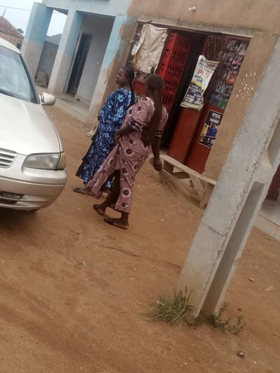 In Russo, Nigeria, two women stand on a dirt road next to a silver sedan. The woman on the left wears a blue and white patterned dress, while the woman on the right is dressed in a pink and white patterned dress and carries a broom. Behind them, a building with a light blue facade features an open doorway leading to a shop selling beverages. A sign advertising "NKU YUANNA PROSION STORE" is visible on the shop. A wooden bench is positioned outside the shop, and a concrete pillar stands in the foreground. The environment is dusty and arid, indicative of a dry climate.