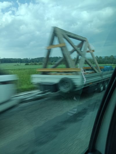 This image captures a blurry, dynamic scene of a trailer carrying a large wooden structure being towed along a road. The trailer is long and flatbed, with multiple wheels visible. The wooden structure appears to be part of a building or a large frame, constructed from thick wooden beams. It is secured to the trailer with visible straps and wooden supports. Underneath the wooden structure, there are also blue tarps visible, suggesting some cargo is covered.
The setting is a rural landscape with a field of green grass and distant trees. The sky is cloudy, with patches of light blue peeking through. The overall impression is of movement, indicated by the motion blur across the entire image. The time of day appears to be daytime, given the ambient light. There are no people clearly visible in the image, only the blur of what might be part of a vehicle on the left.
The image is taken from inside a vehicle, as evidenced by the reflections and the framing of the right edge of the picture, which shows a car's interior. A faint "adidas" logo is visible as a reflection on the surface of the trailer. The location is identified as Šternov, Czechia. The weather seems overcast, contributing to the somewhat muted color palette.