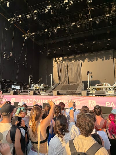 The image depicts a crowd of people gathered in front of a stage at an outdoor event, likely a concert or festival, in Segrate, Italy. The stage area is dark and equipped with numerous lights and scaffolding. In the foreground, the crowd is densely packed, with individuals of various ages and genders. Some are looking towards the stage, while others are interacting with each other. One person in the foreground is holding up their hand, displaying painted fingernails and rings.
A prominent pink banner stretches across the scene, bearing text that appears to be related to the event. Visible portions of the text include "PARCOMUS" and "NO". The time of day appears to be late afternoon or early evening, as evidenced by the warm, golden sunlight casting long shadows. The weather seems pleasant, with clear skies. The overall atmosphere suggests excitement and anticipation for the event. The composition of the image, with the crowd in the foreground and the stage in the background, emphasizes the collective experience of the attendees.