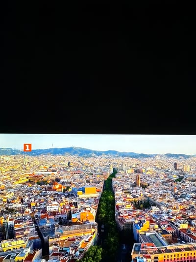 This is an aerial photograph of a densely populated city with a prominent tree-lined avenue dividing it. The buildings are a mix of styles and colors, with many featuring terracotta roofs. In the distance, a range of hills or mountains is visible under a clear sky. On the left side of the frame, a bright orange square with the number "1" in white is overlaid on the image, possibly indicating a point of interest or a marker. The city appears to be bathed in the warm light of either sunrise or sunset. There is no indication of Ognianovo, Bulgaria in this image.