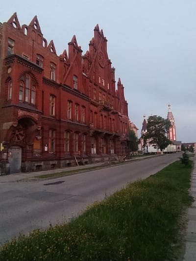 The image captures a street scene in Gusev, Russia, featuring a large, ornate red brick building with Gothic-style architectural elements, including pointed gables and arched windows. To the right of this building, a church with a prominent bell tower and multiple domes is visible. A paved road runs alongside the buildings, with a grassy area and a sidewalk on the right. The sky is overcast. There are no people or vehicles visible in the image.