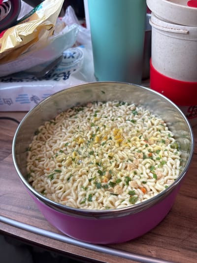 This image displays an indoor scene in the city of Tatarsk, Russia. The central focus is a round, stainless steel bowl with a glossy pink base, resting on a brown, wood-grain surface. The bowl contains instant noodles, which are partially submerged in liquid and topped with green, chive-like pieces, yellow seasoning powder, and small, light-colored dehydrated vegetable or protein fragments. Small bubbles are visible on the surface of the liquid. In the upper left background, a crumpled yellow plastic bag is visible, partially obscuring a white plastic package marked with Russian text, including "TIX XL" and "РОДНОЙ". To the upper right, a light teal-colored cylindrical container, possibly a thermos, stands next to a stack of two beige, lidded containers, with the bottom container featuring a red silicone band. A thin, metallic strip runs horizontally across the lower portion of the wooden surface, beneath the bowl.