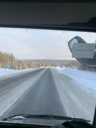 The scene depicts a snow-covered highway in a rural area outside the city of Div'ya, Russia, viewed from inside a vehicle. The asphalt road, partially cleared of snow and ice, extends straight into the distance, flanked by substantial snow banks. Dense evergreen forests, heavily laden with snow, line both sides of the road. The sky is bright and overcast. On the left side of the road, two blue-and-white road signs are visible within the snow-covered verge. On the right side, a small wooden building with a snow-covered roof and other minor structures are visible beyond the snow bank. Within the vehicle's interior, the dashboard and wiper blades are visible at the bottom of the frame. A rearview mirror is partially visible in the upper right quadrant, from which a rectangular tag displaying reversed text, possibly "ЕБОВКУ93," is suspended. A gloved hand is visible in the bottom right corner of the frame.