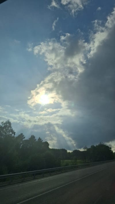 The image depicts a scenic view of a road with a metal guardrail, bordered by lush green trees and foliage on the left. The sky is a prominent feature, with a dramatic interplay of sunlight breaking through dark, heavy clouds, casting distinct rays of light downwards. The sun itself is partially obscured by the clouds, creating a bright, diffused glow. The road surface appears to be asphalt with white lane markings. The overall impression is one of natural beauty and a dynamic sky. The location is identified as Budukan, Russia. There are no people or specific actions depicted in the image.