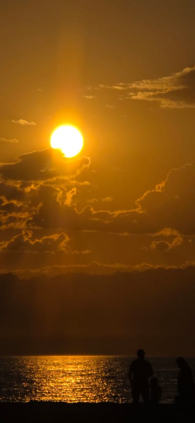 The image captures a sunset over the sea, casting a warm, golden glow. The sun is a bright orb partially obscured by fluffy clouds, creating a dramatic contrast against the orange sky. The sea reflects the sunlight, shimmering with a dappled pattern. In the foreground, silhouetted figures stand on the shore, gazing out at the ocean. The overall scene evokes a sense of tranquility and natural beauty. The location is identified as Basbeshik, Kyrgyzstan, implying a coastal setting, though Kyrgyzstan is a landlocked country and is known for its mountainous terrain. It is possible that "sea" refers to Lake Issyk-Kul, a large saline lake in Kyrgyzstan.