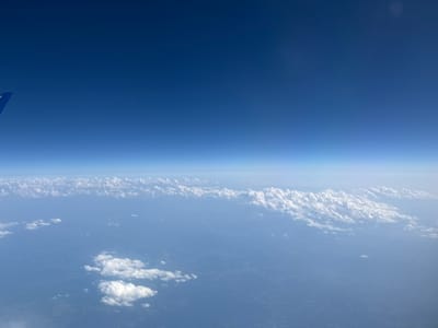 The image is an aerial view from an airplane window, looking down at clouds and the earth below. The sky is a deep, clear blue, with a gradient transitioning to a lighter blue towards the horizon. A small portion of an airplane wing, blue in color, is visible in the upper left corner of the image. Below the sky, a thick layer of white, puffy clouds stretches across the frame, with some areas appearing more dense than others. The ground beneath the clouds is a hazy, muted blue, suggesting land or water partially obscured by atmospheric conditions and distance. There are no visible people, animals, or man-made structures that can be definitively identified. The image does not provide any specific geographical markers to determine its location within Karlovac, Croatia.