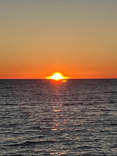 A vertical, medium shot captures a sunset over the Baltic Sea in Meremõisa, Estonia. The sun, a bright, white-hot orb with a distinct halo, is partially submerged on the horizon, casting a warm orange glow across the sky. The sky transitions from a pale grey at the top to a vibrant orange near the horizon. The sea below is dark and choppy, reflecting the sunset with shimmering streaks of light. The horizon line is sharp and distinct, dividing the sky and sea. There are no people, animals, or man-made objects visible in the frame. The overall impression is one of natural beauty and tranquility.