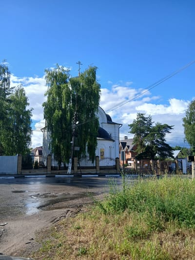 The image shows a bright, sunny day in Novoye, Russia, with a clear blue sky and scattered white clouds. In the foreground, a wet asphalt road with visible potholes and tire tracks leads towards a white building with a black dome and a cross on top, identified as a church by an icon visible on its exterior wall. A black metal fence with stone pillars separates the road from the church grounds. Tall, leafy green trees flank the church and line the fence. Behind the church, several houses with pitched roofs are visible, along with more trees and a power line stretching across the sky. The environment appears to be a semi-urban or suburban area, with elements of both residential buildings and religious structures.
