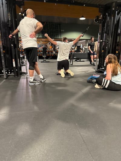 Two men are doing single-arm cable rows in a gym. One man, bald, is facing the cable machine, pulling the cable with his left hand. The other man is turned away from the cable machine, pulling the cable with his right hand. A woman wearing leggings is kneeling in the background. Another person is seated on a bench in the background. Gym equipment is visible in the background.