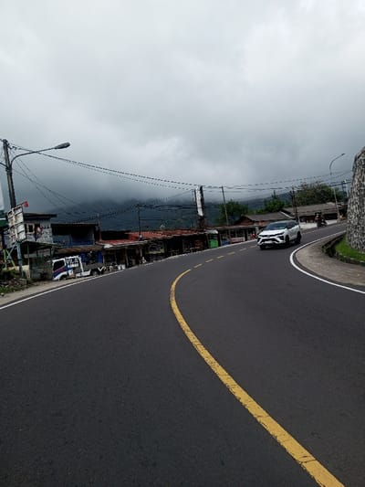 The image displays a curved asphalt road in Saing Ta Mawt Lay, Myanmar, under a heavily overcast sky. A solid yellow line marks the center of the road, which curves to the right. A white Toyota Fortuner SUV is observed in the right lane, moving away from the viewer, while further down, a motorcycle with two riders is also in motion. Along the left side of the road, a series of low-rise, single-story structures, consistent with commercial stalls or shops, are present. A white pickup truck, bearing the text "NILA RAMAD" on its side, is parked parallel to the road in front of these structures. Multiple utility poles supporting numerous electrical wires and at least one streetlight are visible on the left. One person is discernible near the entrance of a structure on the left. The right side of the road is bordered by a low stone retaining wall, behind which a drainage channel is visible. Distant mountains in the background are partially obscured by dense clouds or fog.