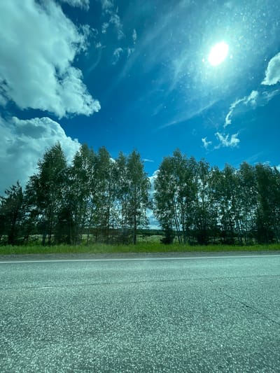 The image shows a road adjacent to a line of trees under a blue sky with clouds and a visible sun. The scene is possibly taken from inside a moving vehicle as indicated by the perspective and angle. The image is rotated. There is no specific information in the image to confirm the location in the city of Средний Постол, Russia.