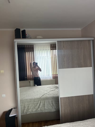 This image depicts an indoor scene, specifically a bedroom, located in Saraia, Bulgaria. The central focus is a full-length mirror, which is a sliding door of a wardrobe, reflecting a male individual and portions of the room.
The person reflected in the mirror is an adult male, shirtless, with a muscular physique, displaying visible abdominal muscles. He is holding a black smartphone with his right hand, positioned to capture his reflection. His left arm is raised, with his hand placed behind his head. He is wearing black athletic shorts that feature a white logo on the left leg, consistent with the "Nike" brand. The individual's facial features are visible through the reflection in the phone screen.
The wardrobe, where the mirror is integrated, consists of multiple sliding doors. The mirror occupies one entire door. Adjacent doors exhibit a two-tone design: an upper section with a greyish-brown wood-grain pattern and a lower section in plain white.
The room reflected in the mirror features light peach-colored walls. A large window dominates the background, fitted with vertical blinds; some are dark brown and opaque, while others are sheer white, allowing diffused light and a blurred view of green foliage outside. Below the window, a white horizontal radiator is mounted. The room's ceiling is white, adorned with a decorative white cornice molding along its perimeter. A large bed is visible, covered with a textured, fluffy, off-white or light grey comforter. The bed frame appears to be upholstered in a light beige fabric. A segment of wooden flooring is visible to the right of the bed. On the left wall, a light-colored rectangular light switch panel is present, along with a white electrical double socket positioned lower on the wall. Adjacent to the socket, a dark piece of clothing and a white notebook or book are resting on a brown upholstered surface, possibly a chair or bench.
On top of the wardrobe, outside the direct reflection of the room, several objects are visible: two dark, rectangular boxes, one bearing the text "CHANEL" and the other "INSOMNIA"; a small, grey, fluffy object; and a light-colored, possibly woven, circular item.
The scene is illuminated by natural daylight entering through the window.