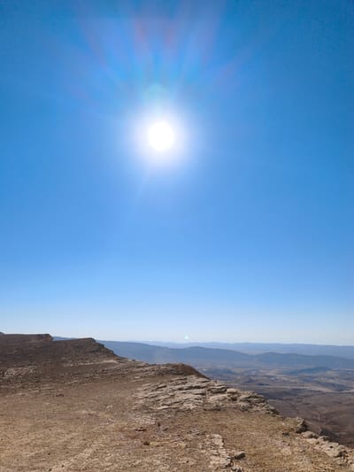 The image depicts a vast, arid landscape under a clear blue sky. The sun is prominent in the upper center of the frame, casting a bright glare and visible rays. In the foreground, a rocky, sandy terrain stretches out, with a distinct cliff edge on the left side of the image. This elevated viewpoint offers a panoramic vista of rolling hills and distant mountains, with a hazy, almost ethereal quality. The landscape suggests a desert environment, likely the Negev Desert, given the context of Yeruham, Israel. There are no people, animals, or man-made structures visible in the immediate foreground or mid-ground, contributing to the sense of natural expanse. The overall impression is one of stark beauty and solitude in a remote, geological setting.