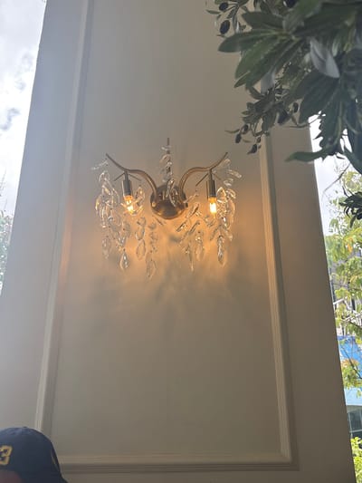 A decorative wall sconce with crystal accents is mounted on a white, paneled wall. The sconce has two visible light bulbs, one of which is illuminated with a warm glow. Branches of an olive tree with dark fruits are visible in the upper right portion of the frame. The bottom left of the image shows the brim of a blue baseball cap with a yellow logo featuring the number "3". The background suggests an outdoor environment with greenery and a cloudy sky. The location is the Mall of Asia complex in Pasay City, Philippines, specifically near the Main Mall's atrium.