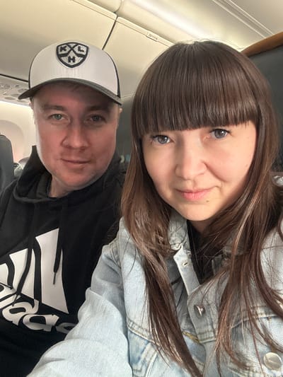 A selfie captures two individuals inside an airplane cabin, with the image rotated diagonally.

The woman is in the upper portion of the frame, featuring long, straight dark brown hair with bangs that cover her forehead. She has captivating blue or grey eyes and a gentle, closed-mouth smile. She is dressed in a light blue denim jacket.

Below and slightly behind her is a man with short, fair hair and blue eyes. He wears a white and black baseball cap adorned with a distinct shield-shaped logo that contains stylized letters, possibly "J," "K," and "H." He is also wearing a black hoodie, on which a large white graphic or text is partially visible on the front. He too has a subtle, content expression.

The background clearly identifies the setting as an airplane interior, showing parts of the overhead compartment and the cabin wall or window. Visible text on the overhead panel includes "28 C," likely indicating a seat or row number. The lighting is typical of an airplane cabin, suggesting artificial light or an overcast day. Both subjects appear relaxed, implying they are travelers on a flight. The provided context indicates the location is Kitsjanovo, Russia.