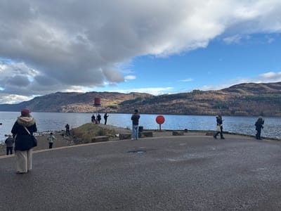 The image depicts a panoramic view of Loch Ness at Fort Augustus, United Kingdom, under a partially cloudy sky with dark grey clouds dominating the left upper frame and clear blue sky on the right. In the foreground, an asphalt-paved area with a yellow double centerline curves towards the right. A rocky shoreline with sparse vegetation borders the paved area to the left, descending into the dark blue loch.

Approximately 12 individuals are present. In the immediate left foreground, a person wearing a dark coat with a fur-lined hood, light trousers, and a red baseball cap stands with their back to the viewer, observing the loch. Along the rocky shore to the left, three more individuals are scattered near the water's edge, one wearing a camouflage-patterned jacket.

A small, elevated landmass protrudes into the loch from the left. A prominent red cylindrical marker buoy with a white base stands centrally on this landmass. Five individuals are gathered on and around this structure, some observing the water, one raising a phone to capture an image. To the right of the paved area, closer to the loch, a red circular marker post is visible. Further to the right, a male individual in a light brown jacket stands facing left. Another individual walks from right to left along the paved edge, holding a phone. At the far right edge of the frame, an additional individual in a dark coat stands, also observing the loch. Distant, treeless hills with brownish vegetation line the far shore of Loch Ness under the horizon.