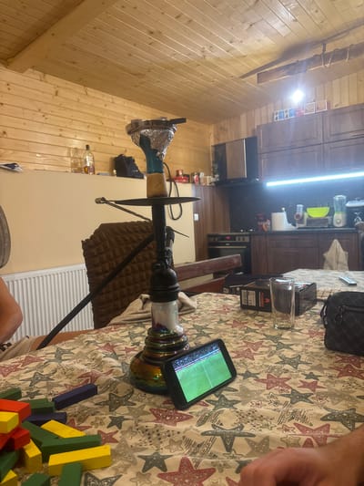 In a room with wooden paneling on the walls and ceiling, a hookah is set up on a table covered with a tablecloth featuring a star pattern. A smartphone is propped up on the table, displaying a soccer game. Nearby, colorful building blocks are scattered. In the background, kitchen cabinets and appliances are visible. The overall scene suggests a casual gathering in a domestic setting.