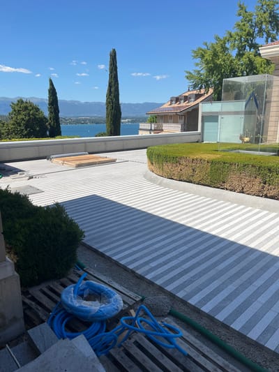 This is a daylight, high-angle, exterior photograph of a modern residential property in Cologny, Switzerland, featuring a panoramic view of Lake Geneva and the surrounding mountains. The foreground is dominated by a newly paved driveway with a striped pattern of light and dark gray granite pavers. To the left, a large green bush is partially visible, with wooden pallets and coiled blue tubing on the ground below. A section of a light-colored stone structure is also visible on the far left.
In the middle ground, the driveway curves around a neatly manicured, low hedge. Beyond the hedge, a glass-enclosed structure, possibly an elevator or staircase, stands next to a building with a tiled roof and dormer windows. The building appears to be a modern villa. The view extends to Lake Geneva, with a distant shoreline and mountain range visible under a clear blue sky with scattered white clouds. Two tall, slender cypress trees stand prominently on the left side of the frame, framing the lake view. The overall impression is of a well-maintained, luxurious property situated in a scenic lakeside location.