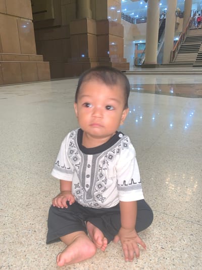 A young child, likely a baby or toddler, is the central subject, sitting on a light-colored, speckled tiled floor. The child wears a white short-sleeved shirt with black and grey geometric patterns and dark-colored pants. With dark hair and dark, inquisitive eyes, the child is looking directly towards the camera.

The setting is a spacious indoor public area, characterized by modern architecture. In the background, tall, light-colored pillars rise, and several flights of stairs or escalators are visible, suggesting a multi-level structure. Reflections of bright, artificial lights are scattered across the polished floor and other surfaces, indicating a well-lit interior. Distant, blurry figures of other people can be seen on upper levels. The environment appears to be a large public building, such as a shopping mall or a transport hub. No visible text is present. The scene takes place in Samarinda, Indonesia.