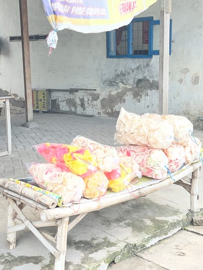 This is a photograph of a food stall in Kepadangan, East Java, Indonesia, selling various types of Indonesian crackers or "krupuk". The stall is set up outdoors, with a rough, grey brick wall forming the background and a concrete ground. A simple wooden and bamboo structure supports the display of goods.

The main subjects are numerous bags of colorful crackers, neatly packaged in clear plastic and tied with string. Some bags contain light-colored, flat crackers, while others hold more vibrant, curled crackers in shades of red, pink, yellow, and white. These are arranged on the bamboo structure, suggesting they are ready for sale. To the left, a large, yellow and purple bag with Indonesian text is partially visible, also tied shut.

In the background, there's a simple building with a weathered, off-white wall. A small, blue-framed window with a grid pattern is a notable detail on this wall. Below the crackers, a yellow sign with black text is visible, which appears to read "RAWON & RUJAK", indicating some of the food items available or related to the stall's offerings.

The lighting suggests it is daytime, with no strong shadows indicating a specific time. The overall atmosphere is that of a casual, local market or roadside vendor. The image is oriented vertically, giving a sense of looking up at the stall.