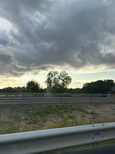 The image depicts a cloudy sky over a rural landscape. In the foreground, a metal guardrail is visible, suggesting the perspective of a vehicle on a road. Beyond the guardrail, a grey asphalt road is present, with a grassy verge and some sparse vegetation. A second, lower guardrail runs parallel to the road. Further in the background, a line of trees forms a dense natural barrier, with a few taller trees interspersed. The sky is dominated by dark, heavy clouds, with patches of lighter sky and some hints of sunlight breaking through near the horizon. The scene appears to be located in Limabacka, Sweden. There are no people or animals visible in the image.