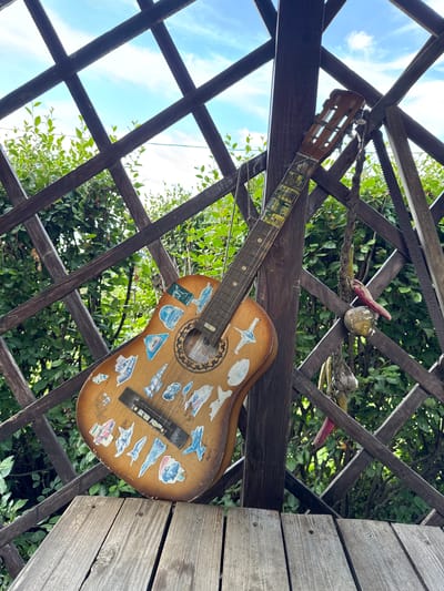 A brown acoustic guitar adorned with various stickers is propped against a dark wooden lattice structure in an outdoor setting. The lattice forms a diamond pattern, with green foliage visible through the openings. A wooden plank runs across the bottom foreground. The sky, visible through the lattice, is blue with scattered clouds. A saw and a rope with dried items hanging from it are also attached to the lattice. The guitar has a worn appearance and features a circular soundhole with intricate detailing. The neck of the guitar has colorful stickers applied to it. The overall scene suggests an outdoor, possibly rustic, environment. The location is specified as Gubarevo, Russia. There are no people present in the image.