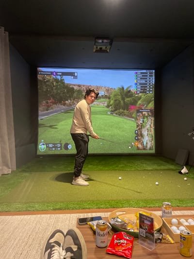 This image depicts an indoor golf simulator bay located in La Purísima, Mexico. A male individual, appearing young, stands center-frame on green artificial turf, positioned to swing a golf club at a white golf ball. He is dressed in a light tan long-sleeved shirt, dark pants, and light-colored sneakers, looking directly towards the viewer. Behind him, a large projection screen displays a virtual golf course with green fairways, palm trees, and distant hills, along with "TRACKMAN" software interface showing player name "SERGIO," scores, and a course map (El Dorado Golf & Beach Club). Overhead, a projector is visible. Multiple golf balls are scattered on the turf. In the immediate foreground, the feet of a second individual wearing dark and white sneakers are visible, resting on a light-colored rug, indicating an observer seated at a low wooden coffee table. This table holds several items: two silver cans of Modelo Especial beer (one open), a red bag of Doritos Nacho chips, a bowl of snacks, a wooden tray with neatly arranged golf balls, and a clear acrylic stand displaying a "GOLF BOX" sign with a QR code. A dark smartphone is also partially visible on the table. The environment features dark walls and ceiling, providing a dedicated space for the simulator.