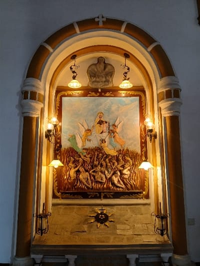 This image presents a religious altar situated within an arched niche inside a building in San Joaquín, Venezuela. The niche features a cream-colored main arch with a mustard yellow trim, supported by two fluted columns. A small cross adorns the apex of the arch.
The central element is a large, multi-tiered relief sculpture. The upper tier depicts a central female figure, likely the Virgin Mary, in a golden gown, holding an infant, surrounded by approximately five winged angelic figures in robes of green, blue, orange, and yellow, against a light blue sky and clouds. The lower tier portrays numerous nude or partially clothed human figures, predominantly male, entangled within detailed orange and brown flames, indicating torment.
The niche is brightly illuminated by two overhead pendant lamps with frosted shades and two wall-mounted lamps with yellow bell-shaped shades positioned on either side of the relief. Below these, two dark metal sconces for candles are present. Beneath the relief is a stone-textured altar or base featuring a central dark, spoked emblem with a cross and central symbols. The surrounding wall is a plain, light cream color.
