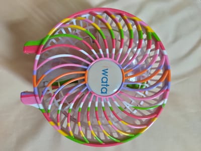 A small, portable fan with a colorful, striped grille is shown from a top-down perspective. The fan's frame and grille are constructed from plastic and are decorated with alternating bands of pink, white, green, yellow, and orange. In the center of the fan, on a circular emblem, the word "wata" is visible in blue lettering. The fan rests on a beige, textured surface that appears to be a fabric, possibly a tablecloth or bedding. The lighting is even, casting no harsh shadows. There are no people or other objects visible in the image, and the background is plain and out of focus. The image's metadata does not provide any information about its location in San Pascual, Philippines.