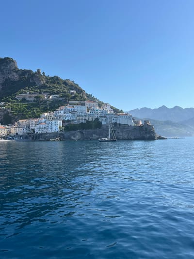 A wide-angle shot captures the Amalfi Coast in Pogerola, Italy, under a clear blue sky. The foreground is dominated by deep blue, rippling water, reflecting the sunlight. In the middle ground, a cluster of white buildings with red-tiled roofs is perched precariously on a rocky cliff face, cascading down towards the sea. A stone bridge with arched openings spans a small inlet near the buildings. A black sailboat with white sails is moored in the water near the cliff. In the background, lush green terraces and more white buildings climb the steep mountainside. Further back, a range of hazy, blue mountains forms the horizon. A few smaller boats are visible in the distance near the shore. The scene evokes a sense of coastal tranquility.