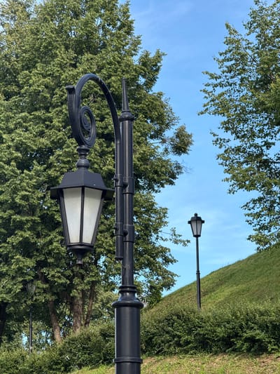 This is a tilted, medium shot of a park scene in Vereya, Russia. The primary subjects are two black lampposts, positioned in the foreground and midground, against a backdrop of lush green trees and a bright blue sky. The lamppost in the foreground is more detailed, featuring an ornate scrolled arm and a white glass lantern. The lamppost in the midground is further away and less detailed, appearing as a simple black pole with a lantern at the top.
The scene is set in a park or a garden, with a well-maintained grassy hill on the right side of the image, also covered with green bushes. The trees are full and leafy, suggesting it is likely summer or late spring. The sky is clear and blue, indicating fair weather. There is no activity or event taking place; it appears to be a still, serene moment captured outdoors.
The time of day is likely daytime, given the bright sunlight illuminating the scene and the clear sky. The lighting is natural and appears to be even, without harsh shadows. No people are visible in the image. There is no visible text in the image. The overall mood is peaceful and tranquil, highlighting the natural beauty of the park.