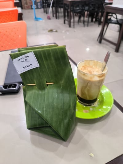An overhead shot captures a dining scene at what appears to be a restaurant or cafe in Sigli, Indonesia. The primary subjects are a packet of food wrapped in banana leaves and a glass of iced coffee. The food packet, secured with a wooden skewer, has a white label with handwritten text that reads "NASI GURUH 'MIMI'" and "DADAR". The glass of iced coffee, served on a bright green saucer, contains layers of dark liquid and frothed milk. A spoon is visible in the glass. The setting is a table with a patterned surface, and the background reveals a dimly lit interior with tables and chairs, suggesting an indoor dining area. Orange plastic chairs are visible in the foreground, and a dark car key or remote is placed on the table near the food packet. The floor in the background is tiled, and a blue mop and broom are leaning against a wall, indicating a cleaning in progress or recent activity.
