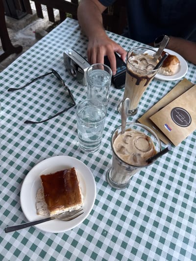 The overhead shot captures a close-up view of a table covered with a green and white checkered tablecloth. Two tall glasses filled with iced coffee and chocolate syrup are prominent, each with a straw and a spoon. To the left, a person's hands are visible, one holding a smartphone and the other resting on the table. Next to the glasses, two empty water glasses and a pair of black sunglasses are laid out. In the upper right corner, a folded brown paper bag with a logo featuring the word "KONDA" in an oval emblem and a small flag is placed on the table. A white plate with a square slice of cake topped with a dark, glossy glaze and a fork rests on the right side of the table. The scene suggests a casual outdoor dining experience, possibly at a cafe or restaurant in Erakovići, Montenegro, given the provided location context. The lighting indicates it is daytime.