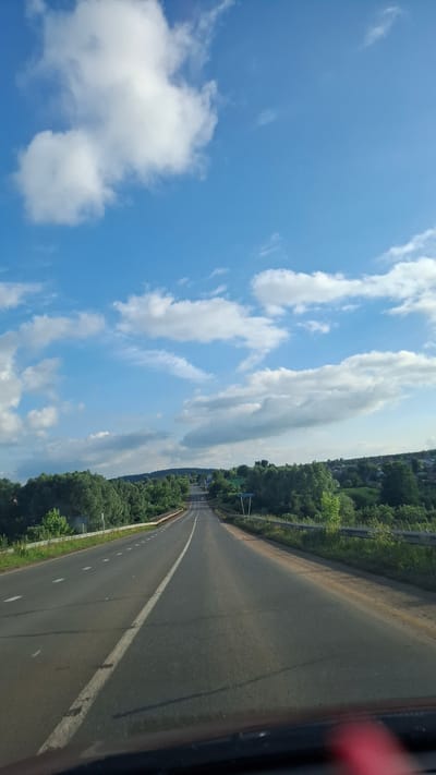 The image depicts a road leading into a rural area under a bright blue sky with scattered white clouds. The road appears to be asphalt with a white dividing line and is flanked by grassy shoulders and metal guardrails. Beyond the road, there are dense green trees on either side, and in the distance, a cluster of houses suggests a village or small town. The perspective is from a vehicle looking forward. The scene appears to be a typical rural landscape in the city of Dokshytsy, Russia.
