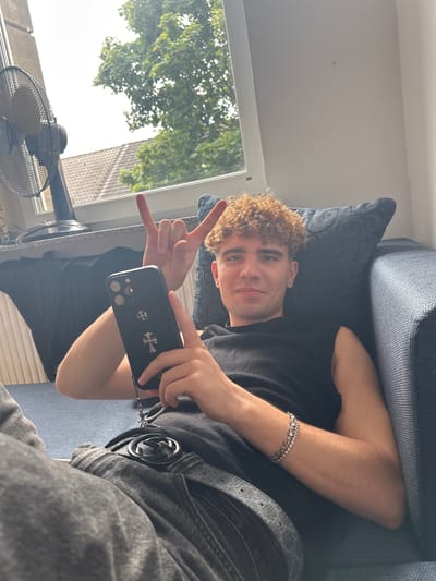 A young Caucasian male with curly, light brown hair is lying on a couch, looking at the camera and holding a black smartphone in his left hand. He is wearing a black sleeveless shirt, gray jeans, and a black belt with a silver buckle. A silver bracelet is visible on his right wrist. He is making a rock-on hand gesture with his right hand. Behind him, a window shows trees and a cloudy sky. A black fan is on a windowsill to the left. The couch is dark blue with a textured pattern. The location is identified as Hamm, Germany.