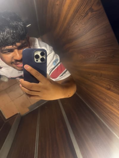 A person with dark curly hair, brown skin, and braces is taking a selfie in an elevator. The person is wearing a white and red jersey. The phone is a blue iPhone with three lenses. The elevator walls are covered in wood paneling. The location is likely Parroquia Ocumare, Venezuela, based on the user's request.