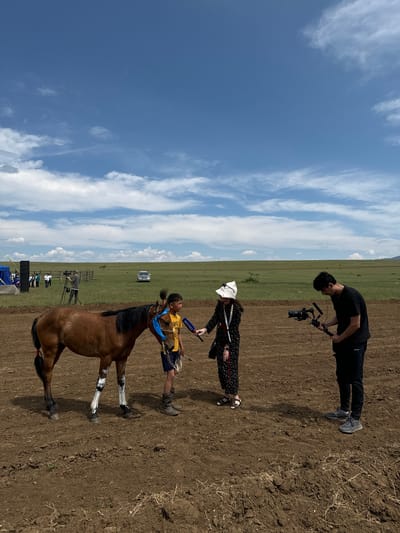 In the city of Orongoi, Russia, on a clear day with scattered clouds, a young boy is being interviewed next to a brown horse. The boy, wearing a yellow shirt and blue shorts, stands to the right of the horse, holding its reins. A woman in a black dress and white hat is interviewing him, holding a microphone. To the right of the woman, a man is filming the scene with a professional camera. In the background, a vast green field stretches to the horizon, with a few trees and a white car visible. A blue tent and a group of people are also present in the distance. The ground in the foreground is bare earth.