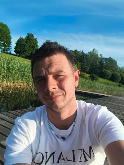 This is a selfie of a man taken outdoors on a sunny day, likely in the late afternoon or early evening given the warm lighting and long shadows. The man is wearing a white t-shirt with a silver chain necklace. He has short, dark hair and a light beard. He appears to be smiling faintly and looking directly at the camera.
The setting is a wooden pier or boardwalk next to a body of water, possibly a lake or river, bordered by tall, green reeds. Behind the reeds, a grassy hillside is visible, dotted with trees and bushes. The sky above is a clear blue with a few wispy white clouds.
The visible text on the man's t-shirt reads "MILANO" in large, black, stylized lettering.
The overall mood is relaxed and serene, suggesting a peaceful moment captured during an outing by the water. The location cues, such as the reeds and the type of vegetation, are consistent with the provided location context of Brocēni, Latvia.