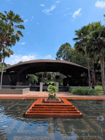In Ban Pong Nuea, Thailand, a wide-angle shot captures a tranquil outdoor scene featuring a modern building with a distinctive curved roof, an elevated water feature, and surrounding lush greenery. The building, constructed with dark framework and a brown tiled roof, has a large open-air entrance, with several cylindrical hanging lamps suspended from the interior. Terracotta-colored steps descend from the building's entrance into a shallow water pool, which is decorated with an intricate pattern of dark stones on its tiled bottom, creating a reflective surface that mirrors the blue sky dotted with white clouds. At the center of the water feature, a small, tiered platform made of the same terracotta tiles supports a cluster of green aquatic plants. To the left and right of the building, tall palm trees with large fronds rise against the clear sky. In the mid-ground, to the right of the building's entrance, a dense patch of tall, feathery green grass grows. A few individuals are visible in the background near the building's entrance, one person walking up the steps and another standing to the far right. The overall environment appears to be well-maintained and serene.