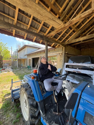 A human is lying on a blue Iseki tractor located in a rural area near a partially constructed brick building and a weathered wooden barn-like structure in Rish, Bulgaria. A black Volkswagen is partially visible, along with construction materials and a white object. The sky is visible behind the structure, and there is green vegetation and trees.