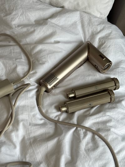 The image depicts a Shark hair styler and two attachments on a white, crumpled bedsheet. The styler, which is a metallic gold color, is positioned diagonally across the frame. The two attachments are located near the styler, and the cord extends from the styler towards the bottom of the frame, terminating in a plug. The scene appears to be indoors, possibly in a bedroom or hotel room. The location is not discernible as Windermere, United Kingdom.