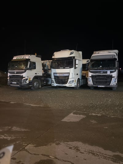 Three heavy-duty trucks are parked side-by-side in an outdoor lot at night in Arad, Romania. The ground, visible in the foreground and to the right, is dark asphalt or concrete and appears wet or damp, possibly from recent rain, with reflections visible on its surface. The background is completely dark.
From left to right (or top to bottom in the original image orientation):
The leftmost truck is a white Volvo FM model. Its license plate, "AR 64 LK", is visible on the front bumper. The "VOLVO" logo and "FM" designation are clearly seen on its grille.
The middle truck is a white DAF XF model. "DAF XF" text is visible on the grille, along with a partially visible company name "P.E.TRANS" (or similar) on the cab side. Its license plate reads "AR 87 ANS".
The rightmost truck is another white Volvo, likely an FMX series due to its rugged appearance, possibly a tipper or construction chassis. "VOLVO" and the horsepower rating "460" are visible on its front. Above the windshield, the text "S.F.T.A" is displayed on the sun visor. This truck also features a yellow rotating beacon on its roof. Its license plate is "AR 38 LK".
All three trucks are white and appear to be commercial vehicles. They are stationary, suggesting they are parked for the night or awaiting deployment. No people are visible. The wet ground and dark surroundings indicate a nighttime scene, possibly after rainfall.