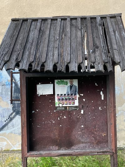 This image shows a close-up, top-down view of a weathered, open wooden structure, possibly a damaged bulletin board or display case, situated outdoors. The left side of the frame features a section of the structure constructed from numerous horizontal, dark gray wooden planks. These planks are distressed, showing signs of age and exposure to the elements, with gaps and splintered edges.
The right side of the frame reveals the interior of the structure, which appears to be a dark, reddish-brown surface with peeling paint and numerous small white fragments scattered across it. A partially visible flyer is affixed to this surface. The flyer has an image of a man in a suit and several small jars or bottles with labels.
Visible text on the flyer includes: "Skoczylas," "w Bielsku-Białej," "PROSTATA," "REKOMENDUJE," "SKLEP ZIELARSKI," "33 812 24 48," and "1 Maja 5, Bielsko-Biała." This suggests the location is in Bielsko-Biała, Poland, and the flyer is advertising a herbal shop.
The background to the left of the wooden structure is a light-colored, textured surface, possibly a wall or pavement. To the right and below, there is green grass and a patch of ground with visible graffiti or painted markings in blue and yellow. The lighting suggests it is daytime, and the overall impression is one of neglect and abandonment, with nature beginning to reclaim the structure.
---
**Description:**
The image captures a distressed, open wooden display or bulletin board located outdoors, with a focus on its aged and damaged components. The left portion of the frame is dominated by a section of dark, weathered wooden planks, exhibiting significant wear and tear, including splintering and gaps. The interior of the structure, on the right, features a dark, reddish-brown surface with peeling paint and scattered debris. A flyer is attached, showing a man in a suit and product images, with text indicating it's an advertisement for a herbal shop in Bielsko-Biała, Poland, with contact information. The surrounding environment includes a light-colored wall or pavement to the left and grass and graffiti-marked ground to the right and bottom. The scene suggests an outdoor location in daylight, conveying a sense of disrepair and the passage of time.