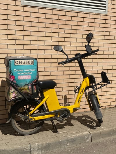 A yellow electric scooter is parked next to a brick wall and a sidewalk in Kotelniki, Russia. The scooter has a black seat and handlebars, and a black carrying case is attached to the back. The carrying case is light blue and has text and images printed on it. The text on the carrying case reads "OH3388" and "Стань частью команды *5580(". The scooter also has a logo on its frame that reads "Яндекс.Банк". The time of day appears to be afternoon, as evidenced by the long shadows cast by the scooter. The weather seems to be sunny and clear. The background consists of a light-colored brick wall and a gray concrete sidewalk.
