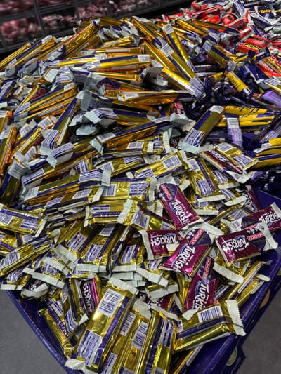 A large pile of individually wrapped chocolate bars is displayed in a dark purple bin, likely in a retail setting. The majority of the bars are golden yellow with purple accents on their wrappers, suggesting they are Cadbury Fry's Turkish Delight. Some of these wrappers also feature a Monopoly branding. The bars are densely packed, with some slightly askew, indicating they have been handled. The background is blurred but appears to be a retail environment with shelves and other products visible, possibly a supermarket in Toormina, Australia. The lighting is bright, highlighting the shiny wrappers.