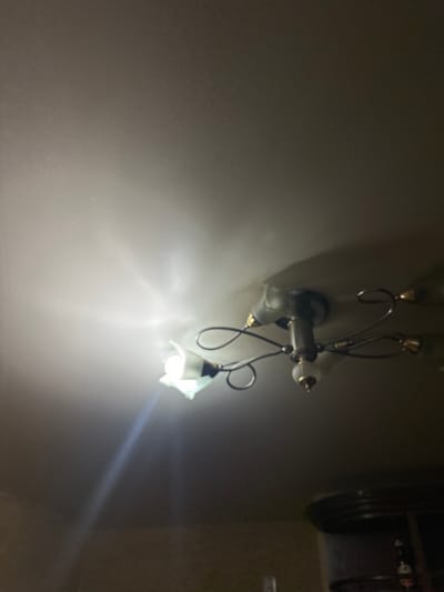 The image shows a close-up of a ceiling with a chandelier. The chandelier has a central white fixture with a gold-colored accent, from which several metallic arms extend, each holding a decorative light fixture with a gold-colored cap and a white glass shade. One of the light bulbs is illuminated, casting a bright light onto the white ceiling. The light creates a hazy glow and some lens flare. The overall environment appears to be indoors, in a room with a neutral-colored ceiling. No people or specific actions are visible. The image does not provide any information that could definitively place it in Aleksandrovka, Russia, or any other specific city.